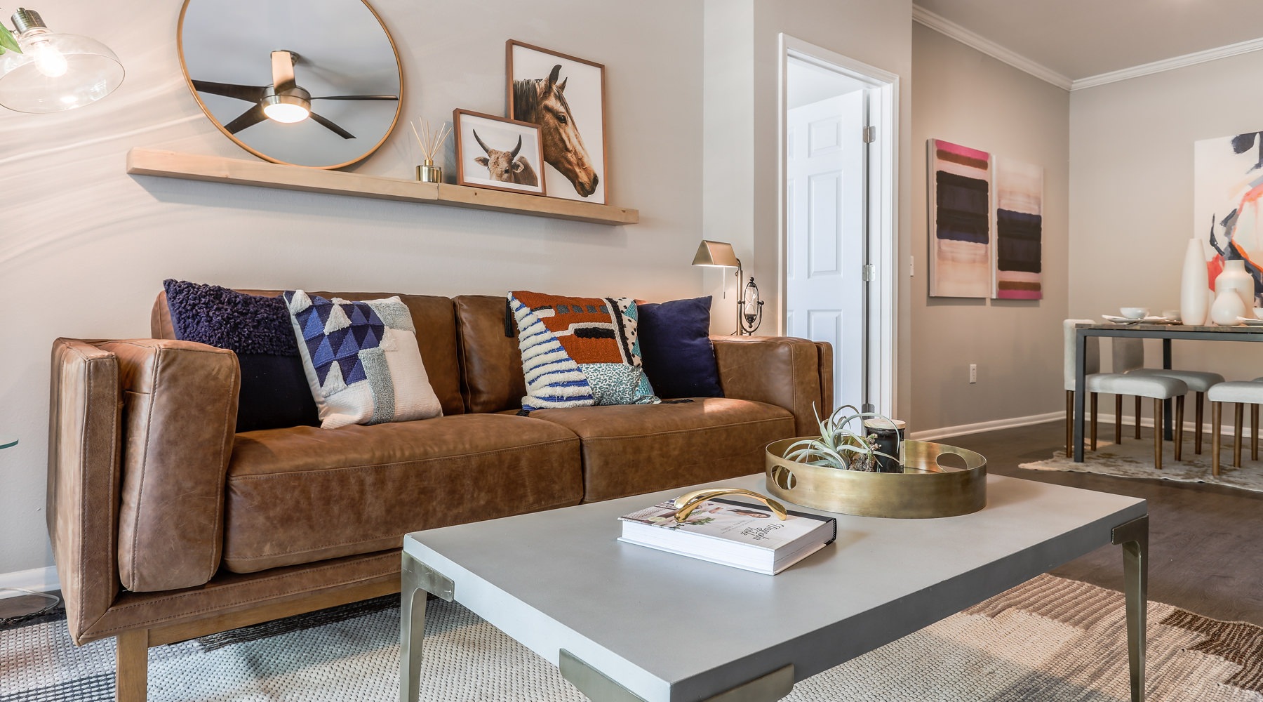 A living room features a brown leather sofa with colorful pillows, a gray coffee table, and a wooden shelf with artwork. A living room features a brown leather sofa with colorful pillows, a gray coffee table, and a wooden shelf with artwork.